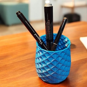 Peut inclure: Un porte-crayons cylindrique bleu texturé avec trois marqueurs noirs à l'intérieur. Les marqueurs ont des étiquettes blanches avec du texte noir. Une étiquette indique "Marqueur Indelebile".