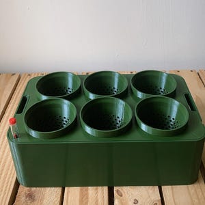 Könnte beinhalten: Ein dunkelgrüner Pflanzkasten mit sechs kreisförmigen Öffnungen. Der Pflanzkasten ist rechteckig mit einem Griff auf einer Seite und einem kleinen roten und transparenten Schlauch auf der anderen Seite. Der Pflanzkasten steht auf einer Holzoberfläche.