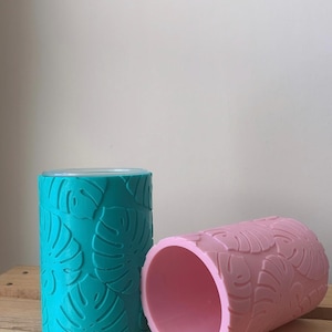 Op de afbeelding: Twee cilindrische drankhouders, één turquoise en één roze, worden getoond. Beide hebben een reliëfpatroon van monstera-bladeren. De turquoise houder heeft een doorzichtige bovenkant. De houders zijn gemaakt van een stevig materiaal en staan op een houten oppervlak.