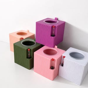 Op de afbeelding: Een verzameling van vijf vierkante, geribbelde zelfbewaterende plantenbakken in paars, olijfgroen, wit en twee tinten roze. Elke plantenbak heeft een ronde opening aan de bovenkant en een verticale waterpeilindicator met een rode dop aan de voorzijde.