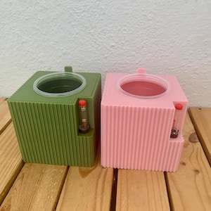 Può includere: Due contenitori quadrati e scanalati, uno verde e uno rosa, sono affiancati. Ognuno ha un'apertura circolare nella parte superiore e un piccolo tubo trasparente con segni rossi sul lato. I contenitori sono su una superficie di legno.