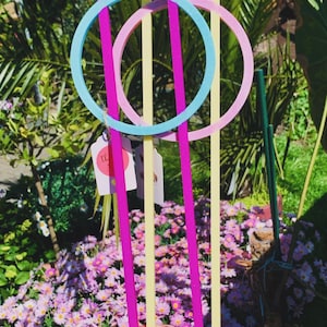 Könnte beinhalten: Eine Hand hält drei farbenfrohe Gartenpflanzenstützen. Die Stützen sind rosa, gelb und blau, mit kreisförmigen und bogenförmigen Designs. Der Hintergrund zeigt einen Garten mit rosa Blumen und grünem Laub.