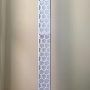 May include: A tall, white, geometric lamp with a honeycomb pattern, standing on a wooden surface. A small, white, rectangular box with a lid is next to the lamp. The lamp is approximately 60 inches tall.