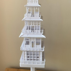 May include: A white, multi-tiered pagoda model with intricate architectural details. The model features multiple levels with overhanging roofs, balconies, and decorative elements. It stands on a white pedestal, showcasing a detailed miniature of a traditional Asian structure.