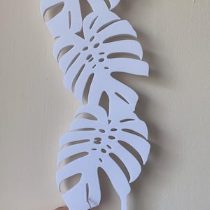 Könnte beinhalten: Weiße, geschichtete Monstera-Blatt-Wanddekoration. Die Blätter sind mit detaillierten Adern ausgeschnitten und haben einen Stiel zum Aufhängen. Ein kleines Etikett mit einer Katzenillustration und den Worten "Thank you" ist angebracht.