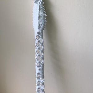 May include: A white garden stake with a detailed, textured design. The top features a dragon-like sculpture, followed by a section with a honeycomb pattern. The stake has a pointed end for easy insertion into the ground.