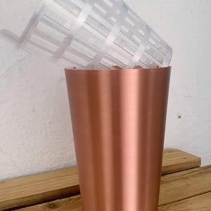 May include: A copper-colored planter pot with a clear, slotted plastic insert. The pot has a smooth, metallic finish and is set on a wooden surface. The plastic insert is designed to hold plants, and the pot is likely for indoor or outdoor use.
