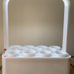 Op de afbeelding: Een wit binnenshuis hydroponisch tuinsysteem met een ingebouwde LED-groeilamp. Het systeem heeft een rechthoekige basis met meerdere cirkelvormige openingen voor planten. Een doorzichtige buis met rode vloeistof staat aan de zijkant.