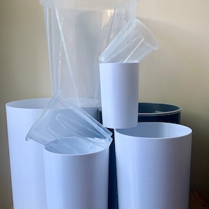 May include: A collection of white and clear plastic containers of various sizes. The assortment includes tall, cylindrical buckets and smaller, cup-like containers, some with lids. The items are arranged on a wooden surface.