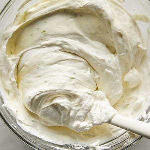 May include: A clear glass bowl filled with a creamy white mixture, with flecks of green. The mixture has a thick, whipped texture. A white spatula with a grey handle is partially submerged in the mixture.