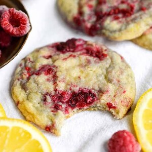 Peut inclure: Un biscuit rond cuit au four avec une surface dorée et des morceaux de framboise rouge visibles incrustés. Le biscuit présente une petite morsure sur le côté droit, révélant un intérieur doux et texturé. Des framboises et citrons sont proches.