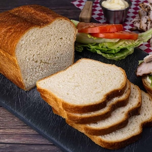 May include: A loaf of freshly baked bread with a golden-brown crust, sliced and ready to be used. Slices of bread are stacked on a black cutting board, next to a sandwich with lettuce and tomato. The bread has a soft, airy interior.