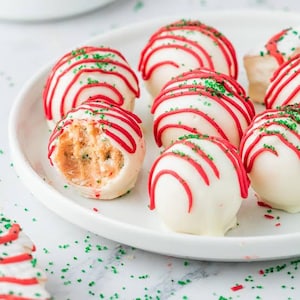Peut inclure: Un assortiment de cake pops ronds enrobés de chocolat blanc lisse, décorés de fins filets de glaçage rouge et de petits cristaux de sucre verts. Un cake pop est entamé, révélant un intérieur de gâteau à la texture claire.