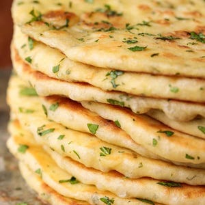 May include: A stack of freshly baked naan bread, golden brown with visible char marks. The flatbread is sprinkled with chopped green herbs, likely parsley or coriander, adding a pop of colour and freshness. The bread appears soft and fluffy.