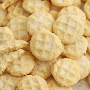 Peut inclure: Gros plan sur un tas de biscuits ronds, jaune clair, avec un motif gaufré et saupoudrés de sucre. Certains biscuits sont cassés, révélant une texture friable. Les biscuits sont disposés de près.