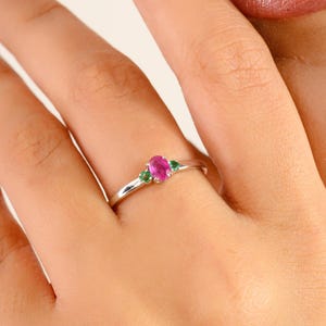 May include: A silver ring featuring a central oval-cut pink gemstone, flanked by two smaller green gemstones. The ring is displayed on a hand with painted nails, showcasing the jewelry's design and color contrast. The ring is a piece of fine jewelry.