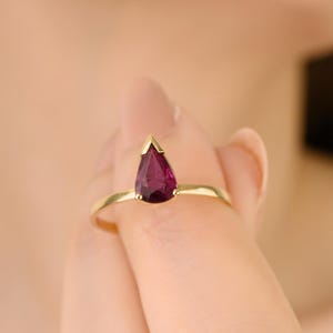 May include: A gold ring featuring a pear-shaped, deep red gemstone. The ring's band is thin and delicate, and the gemstone is set in a gold bezel. The ring is held up in front of a person.