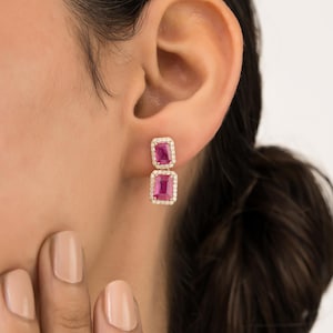 May include: A pair of elegant earrings featuring two rectangular, vibrant pink gemstones. Each gemstone is surrounded by a halo of small, sparkling clear stones, set in a gold-toned metal. The earrings are designed to be worn in the ear.