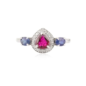 May include: A silver ring featuring a pear-shaped, vibrant pink gemstone surrounded by small, clear stones. Two round, blue gemstones flank the central stone. The ring is set against a white background, highlighting the jewelry's design.