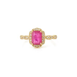 May include: A gold ring featuring a rectangular, bright pink gemstone surrounded by small, clear stones. The ring's band has a delicate, textured design. The ring is set against a white background.