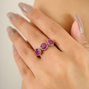May include: A silver ring featuring three round, vibrant red gemstones. The gemstones are set in a row, creating a classic and elegant design. The ring is shown on a hand, highlighting its size and style.