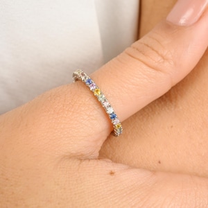 Puede incluir: Un fino anillo de banda de plata con una fila de piedras preciosas de colores. Las piedras preciosas están engastadas en pavé y están dispuestas en un patrón de arcoíris.