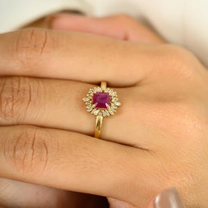 May include: A gold ring featuring a square, deep red gemstone at the center, surrounded by smaller, clear gemstones. The ring's band is a simple, polished gold. The ring is displayed on a person's finger, with the hand in soft focus.