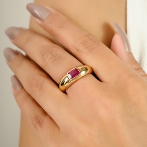 Puede incluir: Un anillo de oro con una gema de rubí rectangular. El anillo tiene una banda lisa y redondeada y un engaste de bisel que sostiene el rubí rojo vibrante. El anillo se muestra en el dedo de una persona.