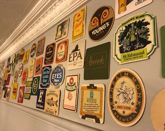 May include: A wall covered in various round and square coasters with different brand names and logos. The coasters are mostly brown, beige, and green. Some of the logos include Harp, St. Edmund, Young's Bitter, and Ephraim Monk Brewery.