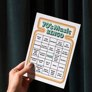 May include: A retro-style bingo card with the text "70's Music BINGO" in a teal and orange design. The card features a grid of song titles from the 1970s. The card is held by a hand against a dark green background.