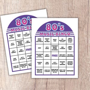 May include: Two 80's Music Bingo cards are displayed on a light wood surface. The cards feature a purple and pink header with the text "80's MUSIC BINGO". Each card has a grid of song titles, including "Hungry Like the Wolf" and "Let's Dance".