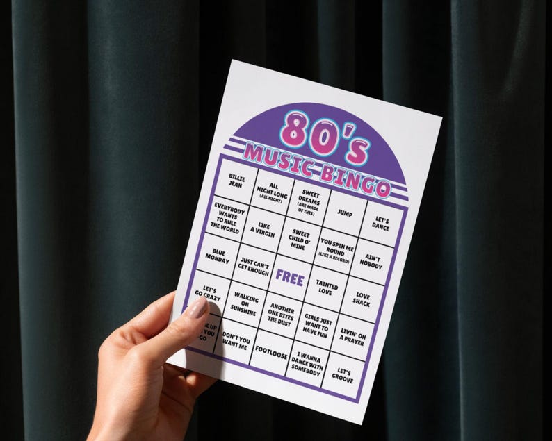 May include: A hand holds a white 80's Music Bingo card with a purple and pink header. The card features song titles like "Sweet Dreams" and "Footloose" in a grid format. The background is a dark teal curtain.