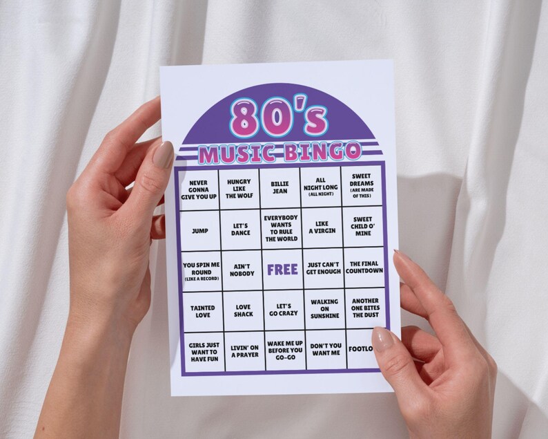 May include: A white 80's Music Bingo card with a purple and pink header. The card features a grid of song titles like "Hungry Like the Wolf" and "Sweet Dreams." The card is held by a person.