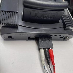May include: A black Atari Jaguar console with a black and silver audio cable connected to the back. The cable has a red and white RCA connector and a black connector labeled "AUDIO-R".