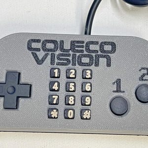 Può includere: Controller ColecoVision grigio con pulsanti neri e un pad direzionale. Il controller ha un tastierino numerico, due pulsanti di azione e il testo "COLECO VISION" stampato in alto.