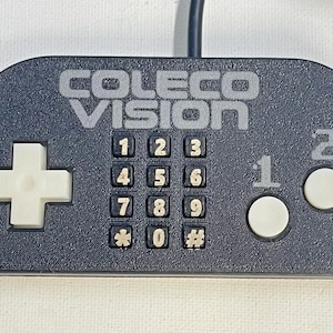 以下が含まれることがあります： 黒いColecoVisionビデオゲームコントローラー。 コントローラーには、方向パッド、テンキー、2つの白いボタン、上部に「COLECO VISION」という文字が印刷されています。