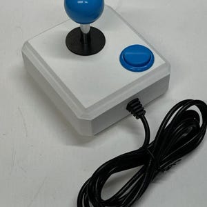 May include: A white arcade joystick with a blue ball-top handle and a blue button. The joystick has a black base and a black cable. The cable is coiled and lies next to the joystick.