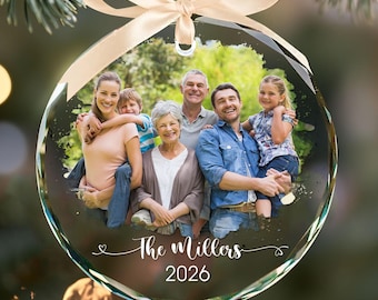 May include: A round, clear glass Christmas ornament featuring a printed family photograph. The ornament is topped with a light beige satin ribbon bow for hanging. The text "The Millers" and "2026" is printed in white script and block font.
