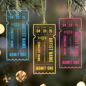 May include: Three rectangular concert ticket ornaments in blue, yellow, and pink, each hanging from a gold string. The tickets feature printed text including SEC 04, ROW 20, SEAT 15, SUN NOV 23 2025, ARTIST NAME, CONCERT NAME, CUSTOM LOCATION, and ADMIT ONE.