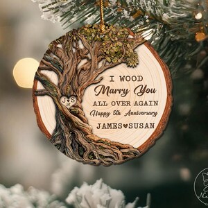 Op de afbeelding: Een houten kerstornament met een boomontwerp en de tekst "I Wood Marry You All Over Again Happy 5th Anniversary JAMES SUSAN". Het ornament heeft een gouden draad om op te hangen.