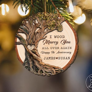 Op de afbeelding: Ronde houten ornament met een boomontwerp en de tekst "I Wood Marry You All Over Again Happy 5th Anniversary JAMES SUSAN". Het ornament heeft een bruine schorsrand en een gouden touwtje om op te hangen.