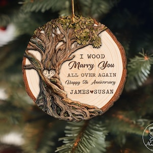 Op de afbeelding: Ronde houten kerstversiering met een boomontwerp. De versiering heeft de tekst "I Wood Marry You All Over Again Happy 5th Anniversary JAMES SUSAN". Opgehangen aan een gouden draad.