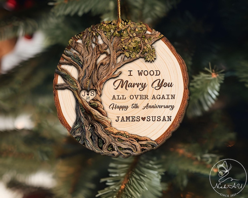 Op de afbeelding: Ronde houten kerstversiering met een boomontwerp. De versiering heeft de tekst "I Wood Marry You All Over Again Happy 5th Anniversary JAMES SUSAN". Opgehangen aan een gouden draad.