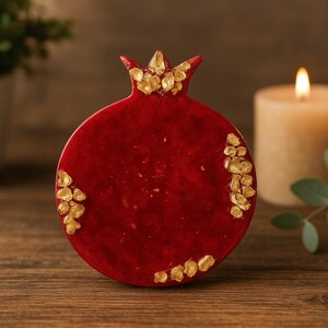 May include: A red pomegranate-shaped decorative object with gold accents. The object has a textured surface and is set on a wooden table. A lit candle and greenery are in the background.