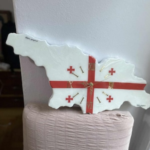 Puede incluir: Un reloj de pared blanco con la forma de Georgia, con un diseño de cruz roja y números romanos dorados para las horas. El reloj tiene manecillas doradas y el texto "Sophia georgiad" está escrito en el reloj.