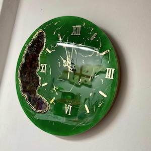 Puede incluir: Reloj de pared redondo verde con números romanos y manecillas doradas. La esfera del reloj tiene un diseño único con una sección de material oscuro y texturizado. El reloj está montado en una pared blanca.