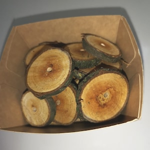 Puede incluir: Primer plano de una pila de pequeñas rodajas de madera redondas con agujeros en el centro. Las rodajas son de color marrón claro y tienen una veta de madera natural. Las rodajas están en un recipiente de papel marrón.