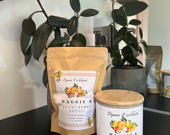 May include: A package and a jar of Maggie's Toilet Bombs, Citrus Blend. The package is tan with a pink stripe and a citrus fruit illustration. The jar is white with a wooden lid and a similar citrus design. The text "Organic & Natural" is above the brand name.