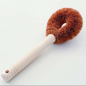 May include: A brown coconut fibre brush with a wooden handle. The brush is shaped like a doughnut and is designed for cleaning dishes.