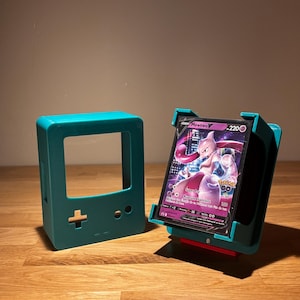 May include: Teal-colored card holder and a matching Game Boy-style display stand. The card holder displays a Pokemon card featuring Mewtwo. The card has a purple and pink design with the number 220.